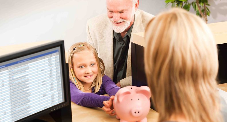 A cheerful grandparent helping a smiling young girl grandchild with her piggy bank savings. Opening a children bank account with the bank teller in a retail bank counter. Photographed indoors in horizontal format.