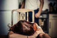 Young woman is reesting her head on a kitchen table, her abusive partner hovering in the background