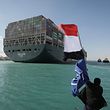 HANDOUT - 29.03.2021, Ägypten, Suez: Ein Arbeiter schwenkt eine Flagge Ägyptens, während im Hintergrund das Containerschiff «Ever Given» in Begleitung von Schleppern über den Suezkanal fährt. Nach tagelanger Blockade durch das riesige Containerschiff ist der Suezkanal wieder frei. Die «Ever Given» wurde am Nachmittag wieder vollständig flottgemacht, wie das Bergungsunternehmen Boskalis mitteilte. Foto: -/Suez Canal Authority/dpa - ACHTUNG: Nur zur redaktionellen Verwendung und nur mit vollständiger Nennung des vorstehenden Credits +++ dpa-Bildfunk +++