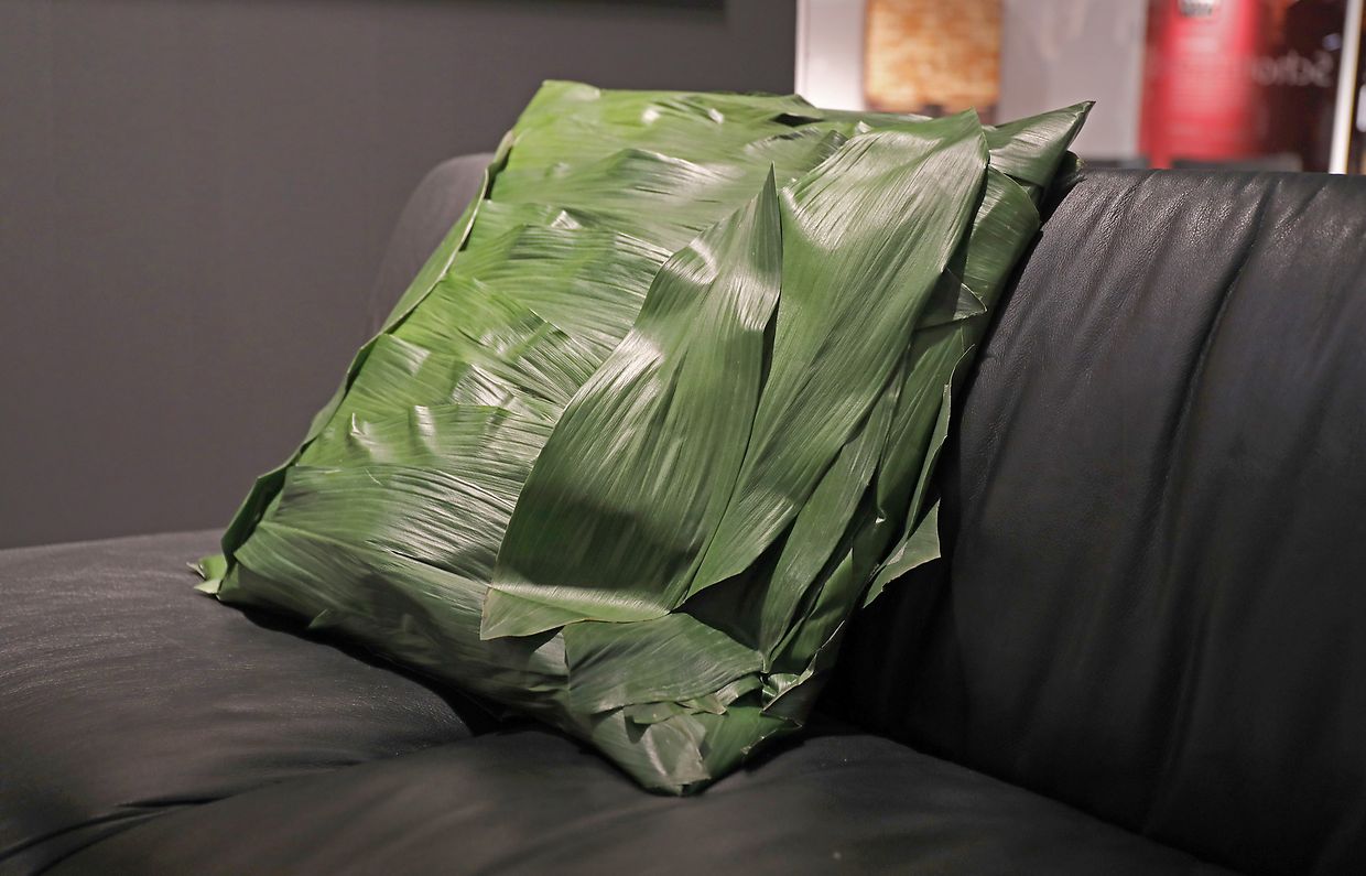 Ein Kissen aus Aspidistra (Schusterpalme) liegt auf einem Sofa am Stand des Herstellers Scholtissek. 