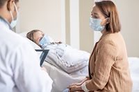 Young brunette woman in protective mask consulting with doctor about her sick friend lying in bed near by in contemporary covid hospital