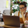 Junger Gärtner, der eine Brille mit Laptop trägt, online mit Kunden im Hausgarten/Gewächshaus kommuniziert, wiederverwendbare Kaffee-/Teetasse auf dem Tisch.