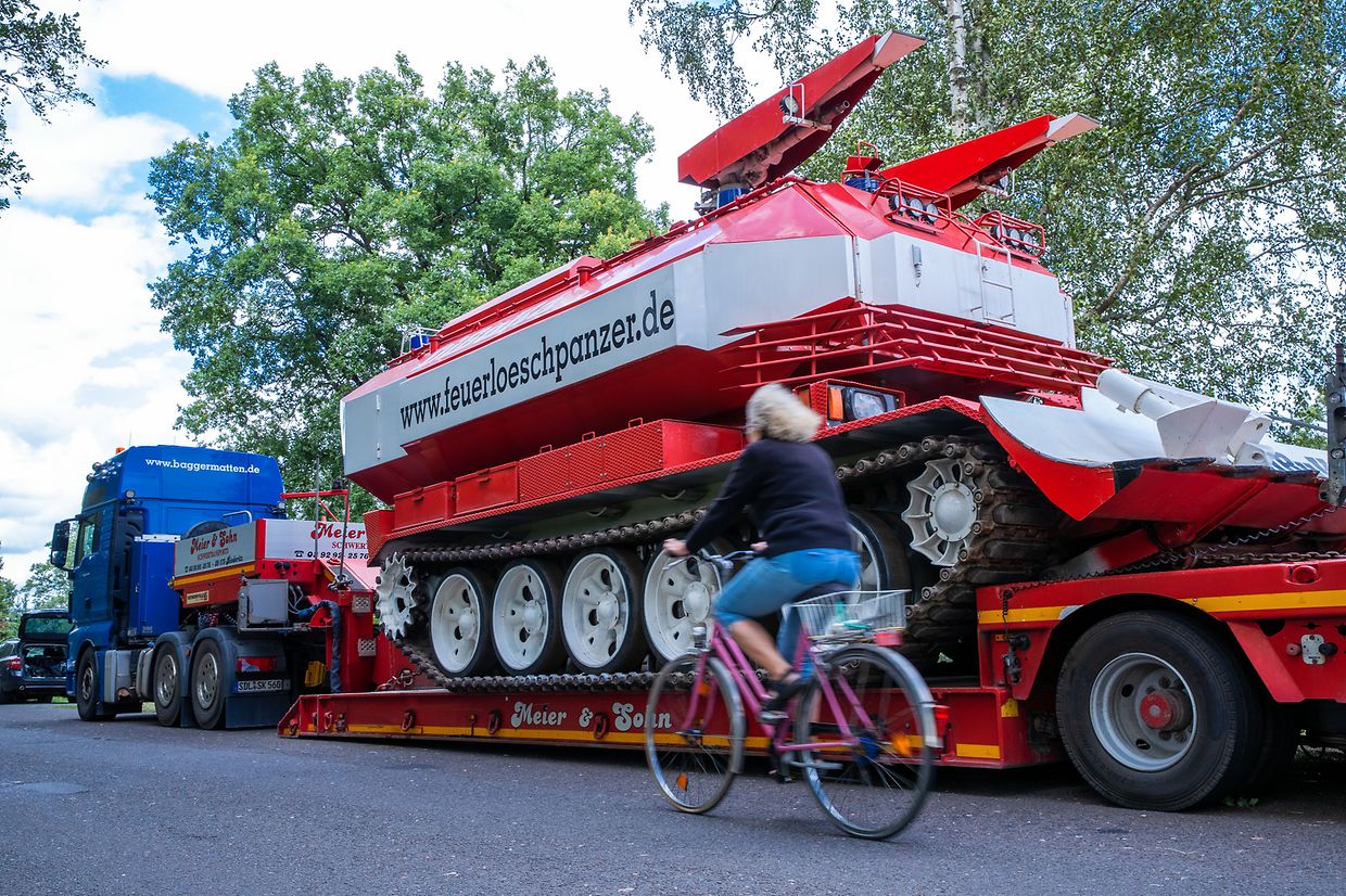 Eine Frau radelt an einem auf dem Tieflader stehenden privaten Feuerlöschpanzer vorbei.