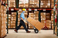 A warehouse worker wearing a protective mask and a hard hat pushes a hand truck and a stack of boxes in a warehouse stacked with inventory.