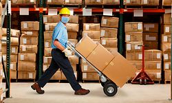 A warehouse worker wearing a protective mask and a hard hat pushes a hand truck and a stack of boxes in a warehouse stacked with inventory.