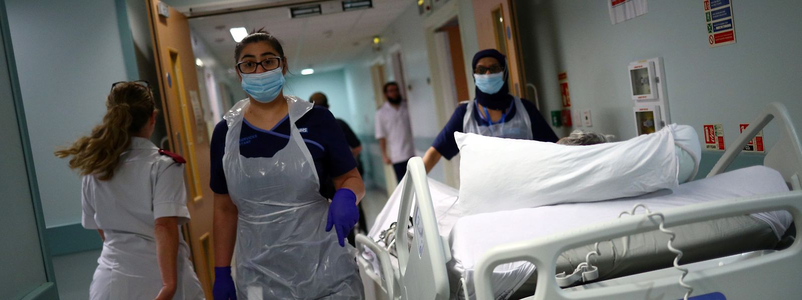 Großbritannien, Blackburn: Medizinische Mitarbeiter des Royal Blackburn Lehrkrankenhauses in Schutzkleidung schieben einen Patienten auf einem Krankenhausbett durch einen Korridor.