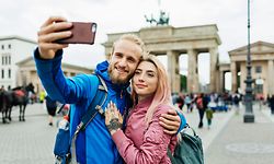A stylish couple embrace to take a selfie in front of local, famous architecture.