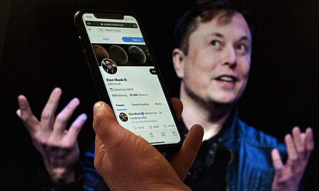 A phone screen displays the Twitter account of Elon Musk with a photo of him shown in the background in a photo illustration produced on 14 April 