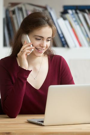 Smiling girl using laptop making answering call sitting at home office desk, happy student talking on phone looking at computer screen, young sales person consulting online customer by cellphone - Image 