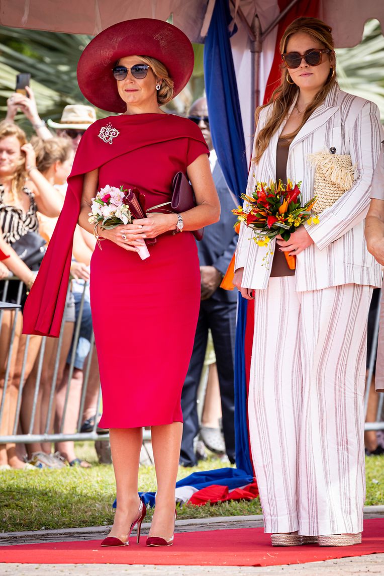 Aruba, 30. Januar:  Anlässlich der offiziellen Willkommenszeremonie auf Aruba war die Garderobe etwas formeller. Königin Maxima bezauberte in einem figurbetonten kirschroten Kleid mit einem für sie typischen Cape-Detail an der Schulter (von Natan Couture, einem der Lieblingsdesigner der Königin). Prinzessin Amalia setzte auf einen leichten Leinenanzug im Streifenlook von Max Mara.
