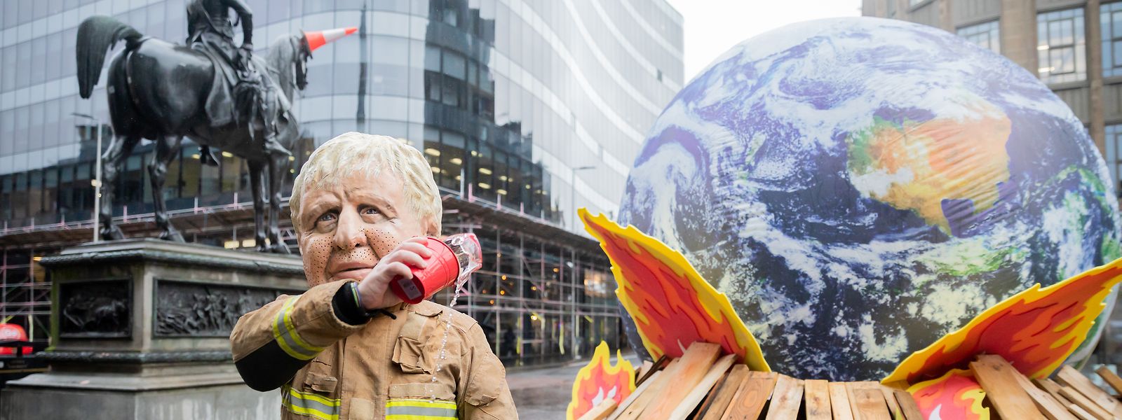 Ein als Großbritanniens Premierminister Boris Johnson verkleideter Aktivist schüttet bei einer Protestaktion von Oxfam in der Rolle eines ineffektiven Feuerwehrmanns Wasser auf einen Globus auf einem Scheiterhaufen.