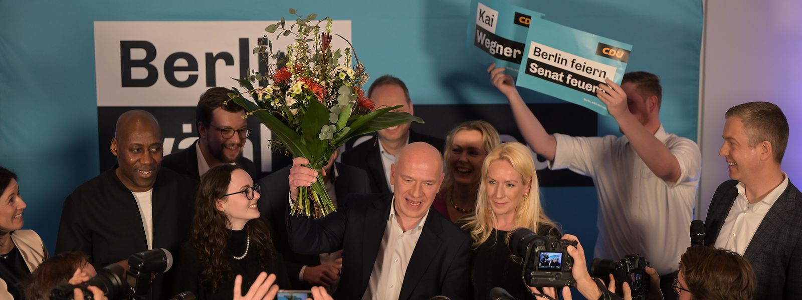 CDU-Spitzenkandidat Kai Wegner steht mit einem Blumenstrauß in der Hand neben seiner Partnerin Kathleen Kantar auf der Bühne der CDU-Wahlparty.