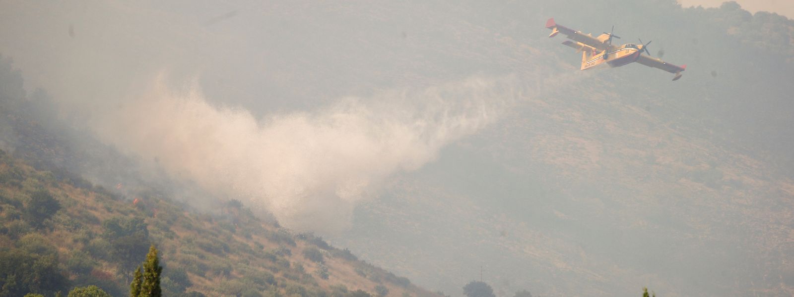 Rauch steigt von einem Feuer im Bergpark Catillo in der Nähe von Tivoli auf, während ein Löschflugzeug darüber Wasser abwirft.