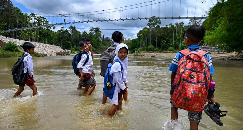 4. Oktober: Mit Schulranzen auf dem Rücken und Schuhen in den Händen überquert eine Gruppe von Jungen und Mädchen einen knietiefen Fluss. Im Hintergrund sieht man die Bauarbeiten an einer Hängebrücke. Bis diese fertig ist, führt der Schulweg für die Kinder aus dem indonesischen Dorf Siron durchs Wasser.