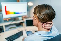 Business young Caucasian woman having neck ache while using computer with charts on display in office, overtime in the office.  Rear view of an attractive professional female Rubbing her neck, working at her home office desk, using a desktop computer and being thoughtful at late night shift while sitting at night