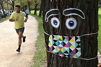 A man jogs past a giant mask and eye display stuck to a tree in Melbourne on May 8, 2020, as Australia's government unveiled a three-stage plan to get the economy back to a new "COVID-safe" normal by the end of July. - The first stage of the plan will allow groups of up to 10 people to gather in cafes and restaurants, for weddings and to take part in outdoor sports. (Photo by William WEST / AFP)