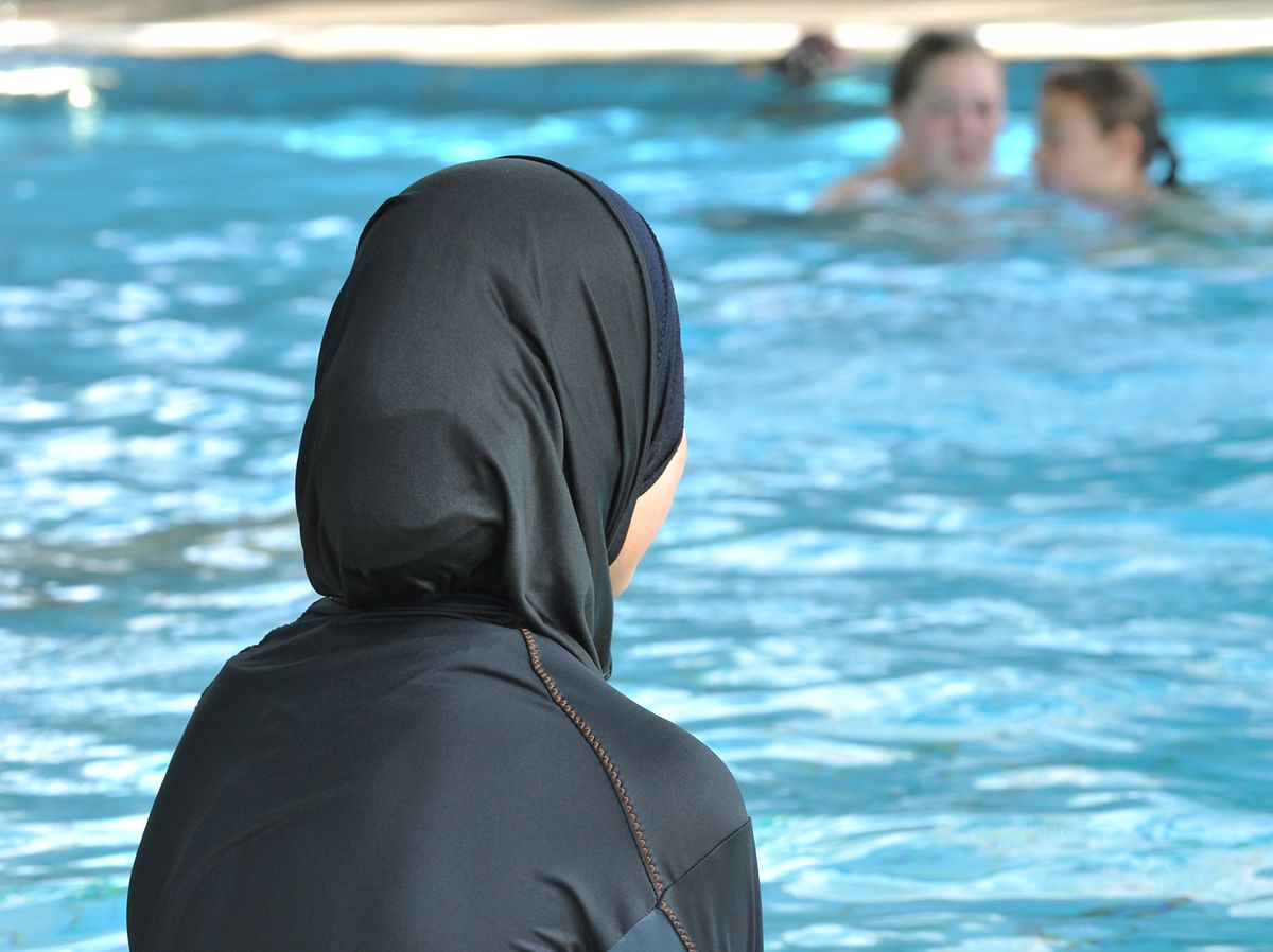 Eine muslimische Schülerin sitzt in einem Ganzkörperbadeanzug (Burkini) am Rande eines Schwimmbeckens. 