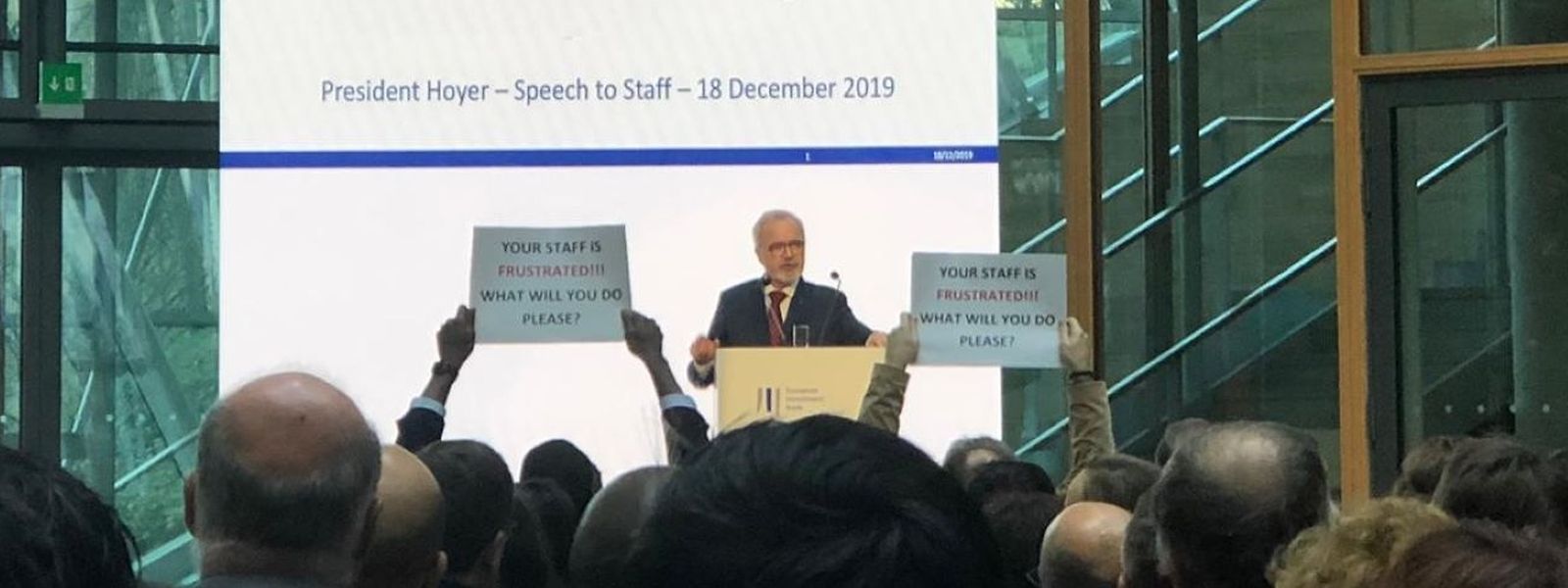 Mitarbeiter halten Schilder hoch, als EIB-Präsident Werner Hoyer 2019 spricht. Dort steht: "Ihre Mitarbeiter sind frustriert! Was werden Sie tun?"
