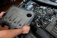  Ein KFZ-Servicetechniker in einer Autowerkstatt hält die Abdeckung vor einem vom Abgas-Skandal betroffenen 2.0l TDI Dieselmotor vom Typ EA189.
